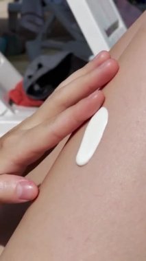 Biri bacağına güneş kremi sürüyor. Kadınların açık tenleri, görünür damarları ve yaraları vardır. Nemlendirici beyaz bir kremdir ve insan eline küçük bir miktar sıkarak kremi sürer. 
