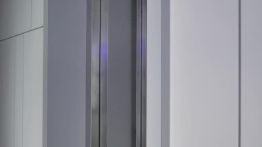 Siyah trençkotlu bir kadın asansör çağırma düğmesine basar ve içeri girer. Bir konut ya da modern bir iş merkezi.