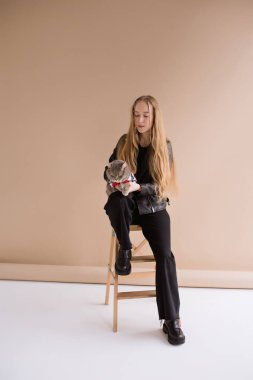 Siyah giysiler içinde gülümseyen, fotoğraf stüdyosunda oturan doğal sarışın bir kadın. Evcil İskoç kedisi olan kız. Dikey portre, 