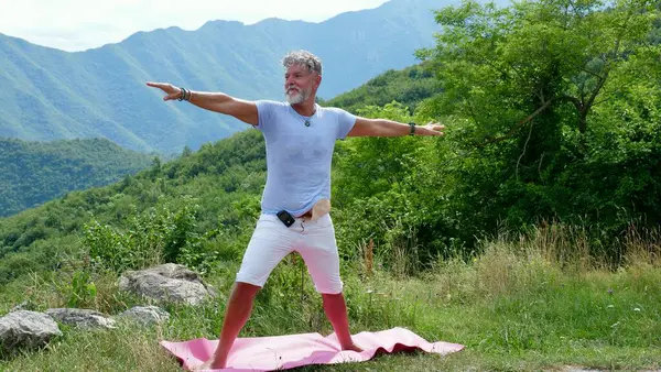 stock image A sick old gray-haired man with diabetes and colon cancer with a colostomy practices yoga in the mountains. Stands in the pose of a warrior