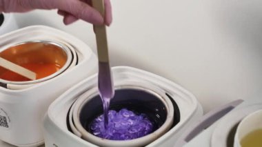 Bir tüy dökücü ustası, mor granüler balmumunu ahşap bir spatulayla eritiyor, yakın plan, yüzü yok.