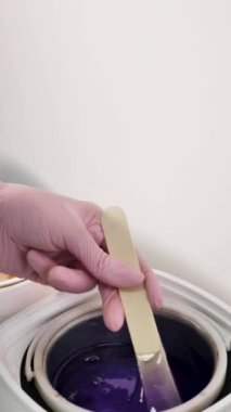 Bir tüy dökücü ustası mor granüler balmumunu ahşap bir spatulayla eritiyor, yakın plan, yüzü yok, dikey.
