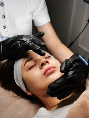 Güzellik uzmanı yüz hücrelerini onarmak için siyah elektrikli eldivenlerde mikro akım uyguluyor. Yakın çekim, dikey