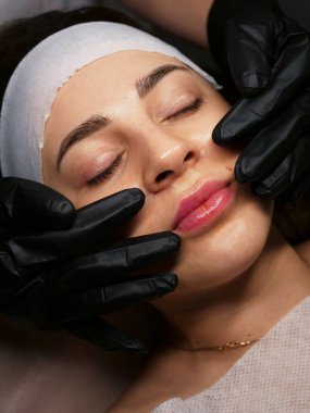 The beautician performs the procedure of microcurrents in black electric gloves to restore facial skin cells. Close-up, vertical clipart