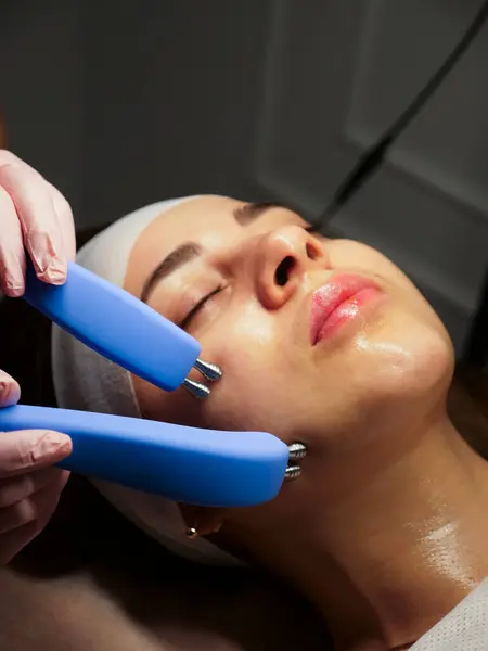 Bir kadın için kozmetik prosedürü. Bir kadın için mikro akım. Güzellik uzmanına bakan bir kadın için. Canlandırıcı etki, yumuşatılmış ten, düzgün ten rengi. Yakın çekim, dikey