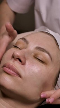 Güzellik uzmanı, krem sürerek nemlendirici sürüyor ve müşterilerin yüzüne masaj yapıyor ve salonda kırışıklık giderici masaj yapıyor. Dikey, yakın plan