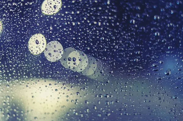 Rainy window at night with water drops and blurry city lights.