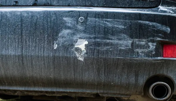 Close up of a dirty car bumper with visible damage including deep scratches, scrapes, and a peeled area revealing the underlying material.