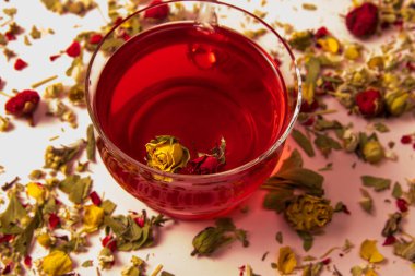 Arka planda gül goncası ve bitki ve çiçek çayı olan cam bir fincanda çay.