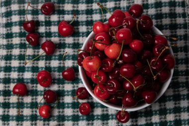 Yeşil ve beyaz kareli mutfak bezinde beyaz bir kasede tatlı kirazlar.