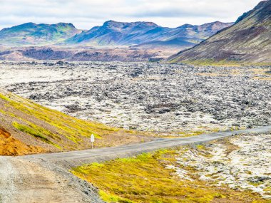 A road winds beside a vast, solidified lava field under a cloudy sky, with rugged hills in the background, depicting the stark terrain of Reykjanes Peninsula in Iceland. clipart