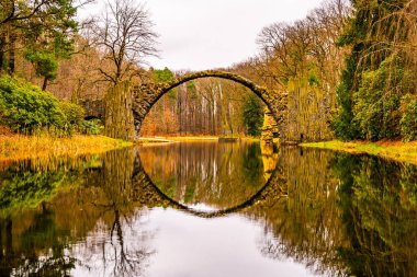 The Rakotzbrucke, also known as Devils Bridge, is reflected in calm waters on an overcast autumn day, surrounded by vibrant foliage. Germany clipart