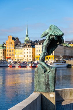 Wings - a bronze sculpture stands at Skeppsholmen Bridge with a view of colorful buildings and boats along Stockholm waterfront under a clear blue sky. Stockholm, Sweden clipart