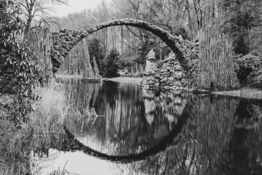 Rakotz Bridge, German: Rakotzbrucke, forms a perfect stone arch reflected in the calm waters of a serene German lake, surrounded by bare trees. Black and white image. clipart