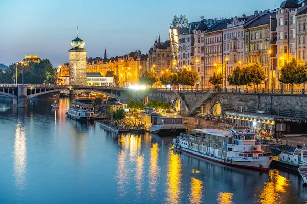 stock image Sitkov Water Tower and the Vltava Embankment in Prague, Czechia. The historic water tower stands tall against a backdrop of brightly lit buildings lining the riverbank. Prague dusk picturesque