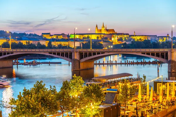 stock image A view of Prague Castle from the Vltava River embankment at dusk, with a bridge spanning the river and a vibrant cityscape in the background. Illuminated by golden lights, creating a warm atmosphere.