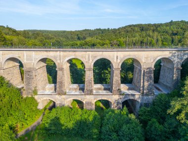 The Sychrov Railway Bridge rises gracefully above lush greenery, showcasing its impressive arches against a backdrop of vibrant trees. clipart