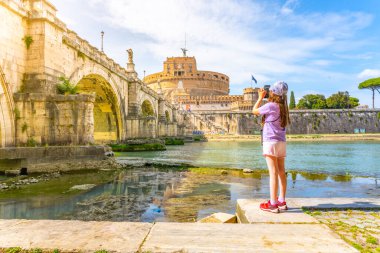 A young girl captures a photo of Castel SantAngelo and a bridge in Rome with her smartphone on a bright sunny day. Rome, Italy clipart