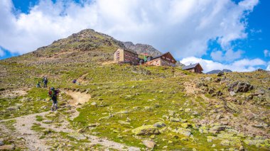 Hikers trek along a path towards Breslauer Hut, nestled in the stunning Otztal Alps of Austria, surrounded by rocky terrain and lush greenery under a bright blue sky with scattered clouds. clipart