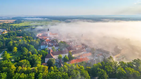 stock image Morning fog blankets Trebon, creating a mystical atmosphere over the landscape. The aerial view reveals the towns architecture and lush greenery, showcasing the serene beauty of Czechia at dawn.