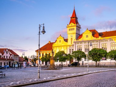 The historic Town Hall stands proud in Marianske Square at dusk, illuminated by warm evening light. Nearby, charming buildings and vibrant greenery create a picturesque atmosphere. clipart