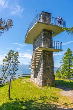 A visitor explores the Gaflei lookout tower in Liechtenstein, enjoying panoramic views of the surrounding mountains and landscape under a clear blue sky. clipart