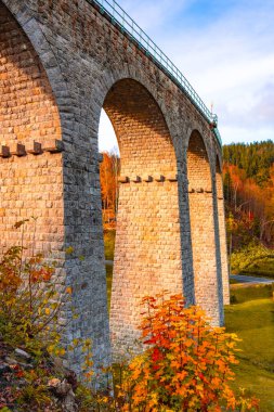 On a sunny autumn day, the railway viaduct in Smrzovka showcases stunning stone arches surrounded by vibrant fall foliage, creating a picturesque landscape in the Jizera Mountains of Czechia. clipart