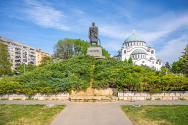 A prominent monument of Karadorde stands proudly in the foreground, framed by lush greenery and the stunning Saint Sava Cathedral in the background, under a clear blue sky. clipart