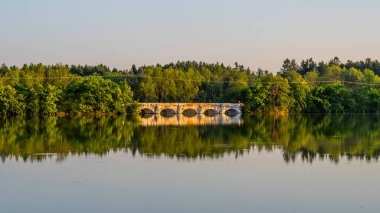 The stone inundation bridge, dating back to the 18th century, gracefully spans Vitek Pond, reflecting the serene evening light among lush greenery in South Bohemia. clipart