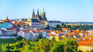 The grand architecture of Prague Castle towers above the vibrant rooftops of the city. Lush greenery complements the lively urban landscape, creating a picturesque view on a clear day. clipart