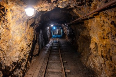 Deep within the Jachymov mine shaft in Czechia, a narrow tunnel reveals old mining tracks and ambient lights illuminating the rocky walls. This location holds a rich history of mineral extraction. clipart