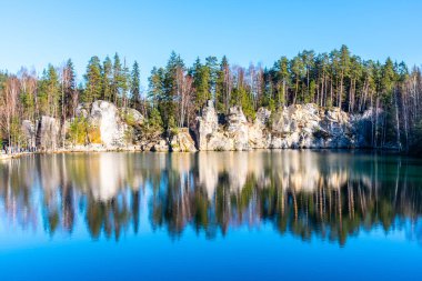 Piskovna Lake in Adrspach Rocks showcases stunning natural beauty. Surrounded by tall trees and rock formations, the calm water reflects the serene landscape on a clear day. clipart