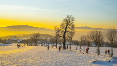 At sunset in Prichovice, Czechia, people gather in the snowy landscape. The golden light casts a warm glow over the peaceful hills and silhouettes of trees, creating a serene atmosphere. clipart