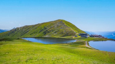 The Wildkogel water reservoir reflects the clear blue sky, surrounded by lush green hills and alpine scenery in the Austrian Alps, creating a serene environment for visitors and nature enthusiasts. clipart