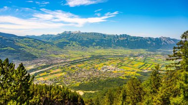 A stunning vista from Gaflei lookout tower showcases the picturesque town of Schaan, surrounded by lush greenery and mountain ranges under a clear blue sky. clipart