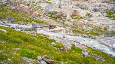 A wooden bridge spans a rushing stream sourced from the Schlaten Glacier, surrounded by rocky terrain and vibrant green vegetation in Hohe Tauern National Park. clipart