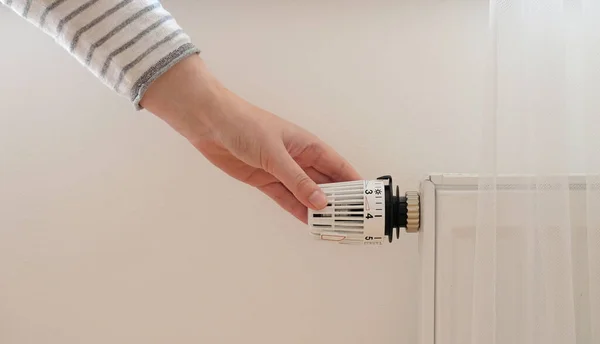 Woman regulates temperature at home with the heating thermostat to save energy, close-up with hand. Temperature control valve on radiator. White heater on wall with curtain in modern apartment.