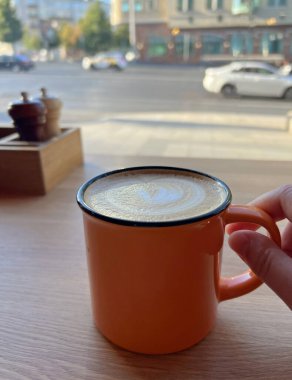 Turuncu seramik kapuçino bardağı ya da köpükle süslenmiş latte. Sokağa, yola ve hareket halindeki arabalara bakıyor. Sabah kahvesi. Sıcak Kafe 'de Sıcak Kahve Zamanı