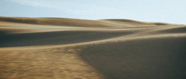 Kurak bir çöl bölgesinde yer alan geniş kum tepecikleri, güzel, berrak bir mavi gökyüzü ile betimlenir.