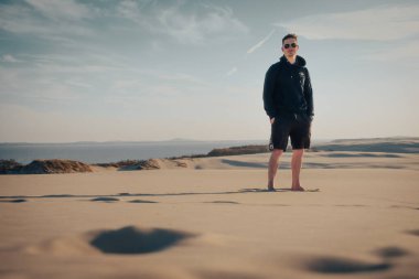 Bir adam uzun bir kum tepesinin üzerinde duruyor, siyah kapüşonlu ve şortlu, çölün geniş manzarasıyla çevrili.