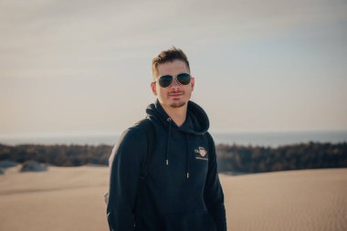 Bir adam uzun bir kum tepesinin üzerinde duruyor, siyah kapüşonlu ve şortlu, çölün geniş manzarasıyla çevrili.