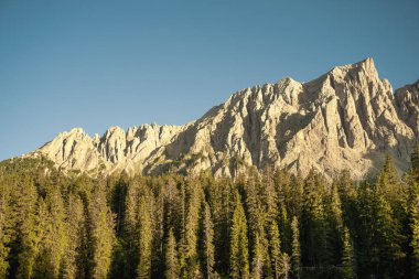 Yüksek bir dağ, canlı yeşil ağaçlarla kaplı, arkasında sonsuz uzanan parlak mavi bir gökyüzüne karşı görkemli bir şekilde duruyor.