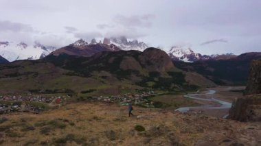 Turuncu sırt çantalı gezgin Hiker Adam Clouds ve El Chalten Town 'daki Fitz Roy Dağı' na bakıyor. Tepeler ve Dağlar. And Dağları, Patagonya, Arjantin. Hava görüntüsü. İnsansız Hava Aracı Yan Uçuyor