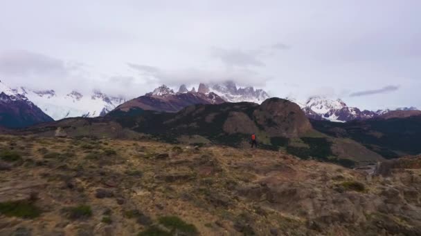 オレンジのリュックを持つ男立ち 雲の中にマウントフィッツロイを見て チャルテン町 丘陵と雪に覆われた山々 アンデス パタゴニア アルゼンチン 空中展望 ドローンは低レベルで前方に飛ぶ — ストック動画