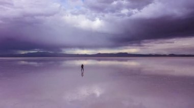 Adam ayakta duruyor ve Uyuni Tuz Düzlükleri 'ne bakıyor. Hava görüntüsü. Altiplano, Bolivya. Yağmur mevsimi. Suyun yansıması. Drone İleri Uçuyor. Geniş açı. Dramatik ve Fırtına Bulutları. Solo Seyahat Konsepti