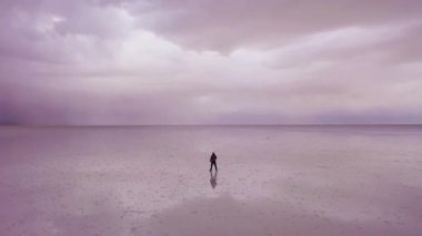 Yalnız turist ayakta duruyor ve Uyuni Tuz Düzlüklerine bakıyor. Hava görüntüsü. Altiplano, Bolivya. Yağmur mevsimi. Suyun yansıması. Yörüngede. Geniş açı. Dramatik ve Fırtına Bulutları. Solo Seyahat