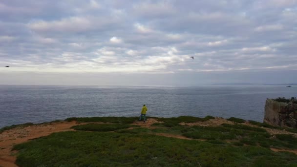 黄色いジャケットのロンリーマンは クリフの端に立って 曇りの日に大西洋を見ています ペニーチェ ポルトランド アリアルビュー ドローン フライ フォワード — ストック動画