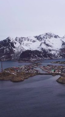 Kışın Henningsvaer Balıkçı Köyü ve Festvagtind Dağı. Norveç Denizi. Lofoten Adaları, Norveç manzarası. Hava görüntüsü. Drone Yana ve Yukarı Uçuyor. Dikey Video