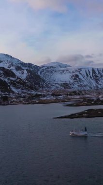 Fjord 'da küçük bir gemi, Fredvang Village ve Sunset' te kışın dağlarda. Lofoten Adaları, Norveç manzarası. Hava görüntüsü. İnsansız hava aracı yandan uçuyor. Dikey Video