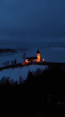 St. Primoz Kilisesi ve Felicijan Kış Akşamı Sisli Alacakaranlığı 'nda aydınlatılmış. Bulutlu Gökyüzü. Jamnik, Slovenya. Hava görüntüsü. Yörüngede. Dikey Video